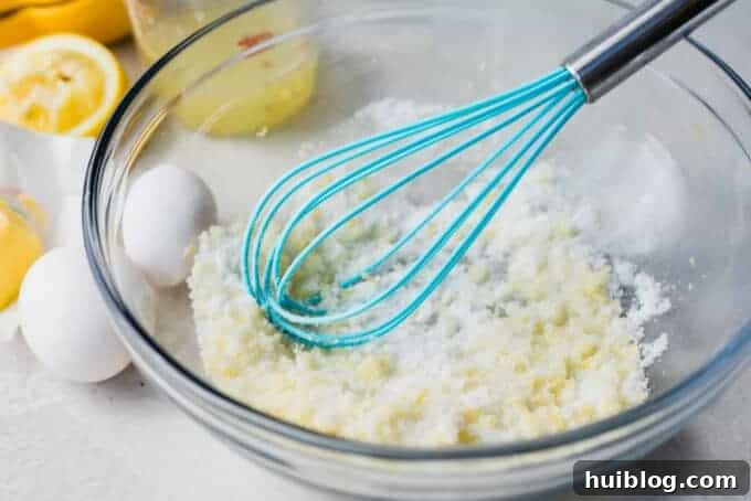 Glass bowl filled with sugar and lemon zest with eggs on the side and a blue whisk.