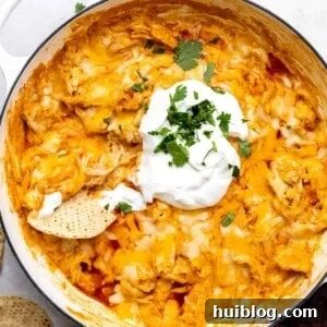 White skillet filled with cheesy chicken enchilada dip, topped with sour cream and with a tortilla chip stuck in it.