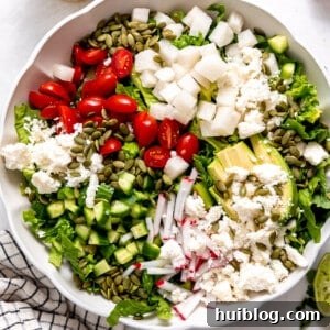White bowl filled with chopped romaine lettuce topped with tomato, avocado, radishes, cucumber, cotija and pepitas.