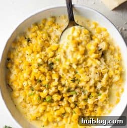 White bowl filled with creamy homemade creamed corn, accompanied by a serving spoon and a wedge of parmesan cheese.