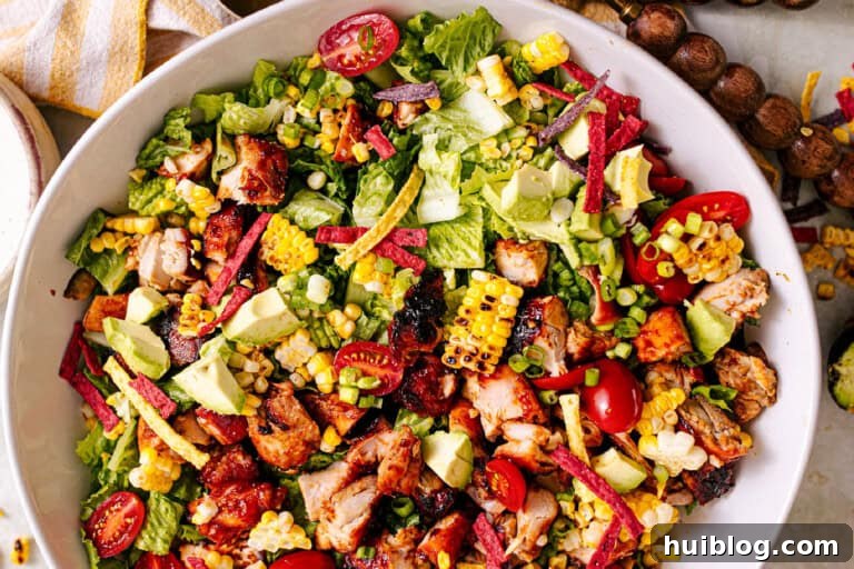 Close-up of a vibrant BBQ Chicken Salad in the process of being assembled, showing layers of fresh ingredients like romaine, chicken, corn, tomatoes, avocado, and green onion.