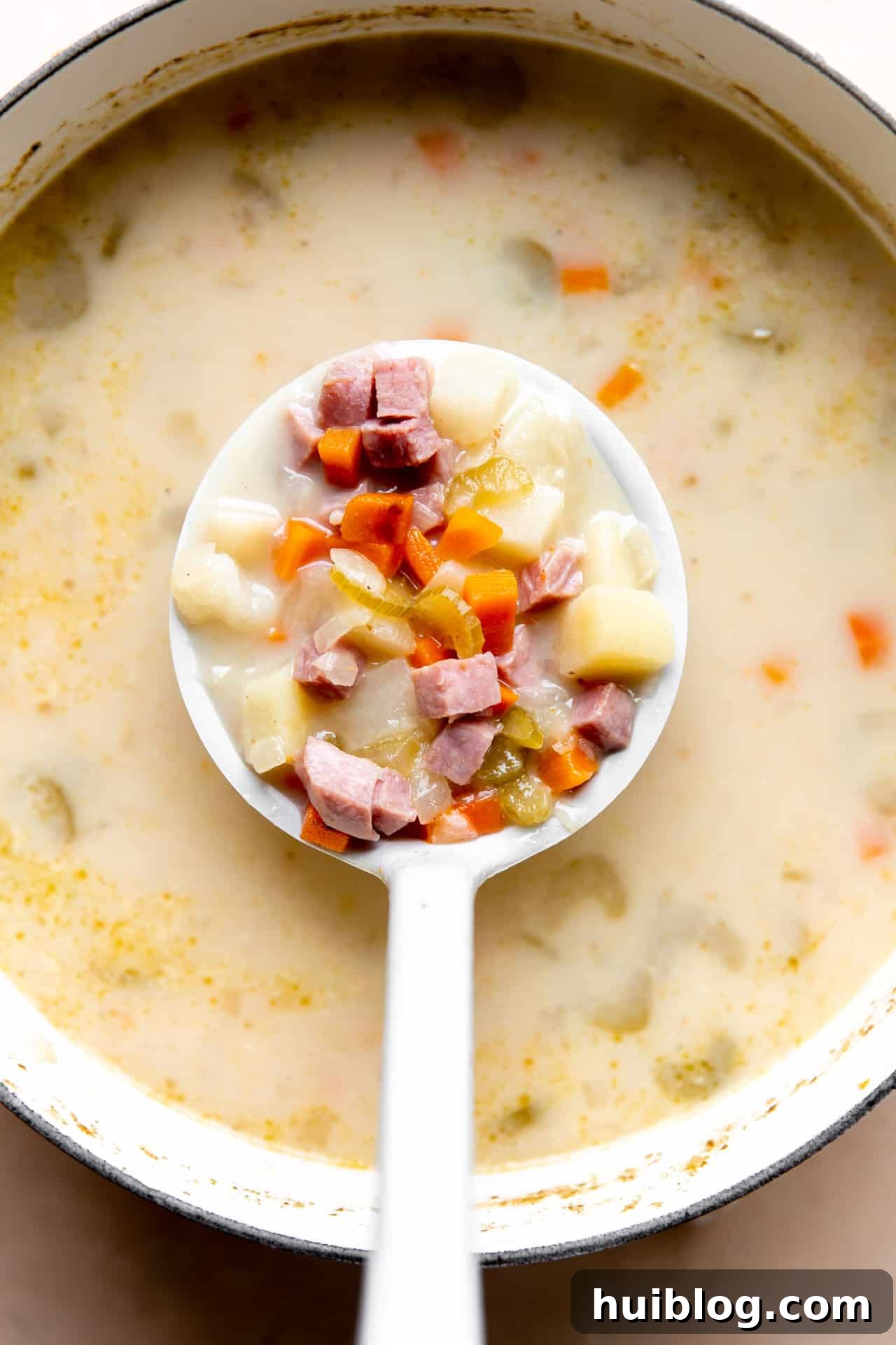 A large spoon scooping up a generous portion of ham bone soup, revealing succulent pieces of ham, tender potatoes, and vibrant vegetables submerged in a rich, creamy broth.