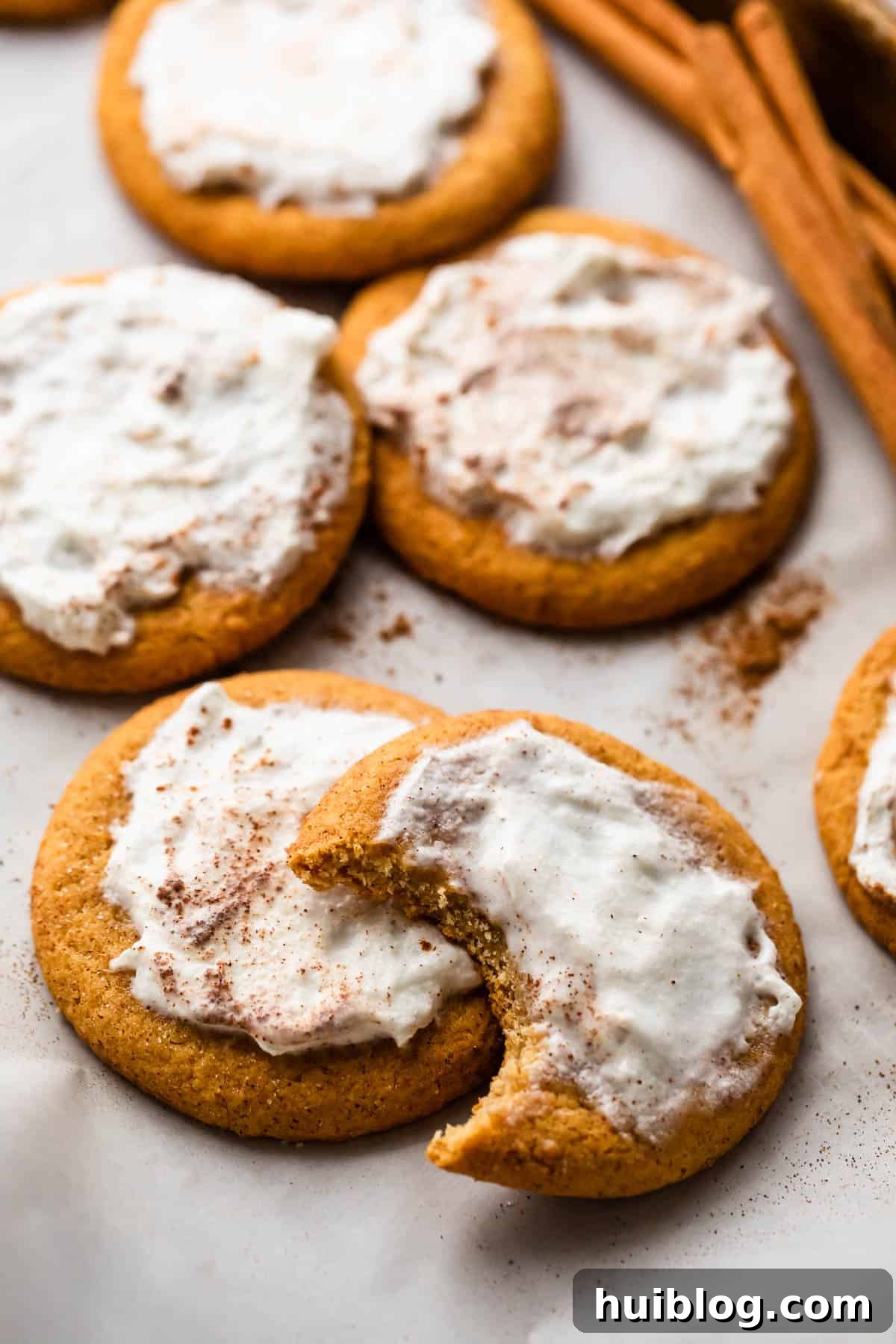 Frosted Pumpkin Cookies arranged on a white background with one cookie halfway bitten off and a few cinnamon sticks set at the top of the right corner.