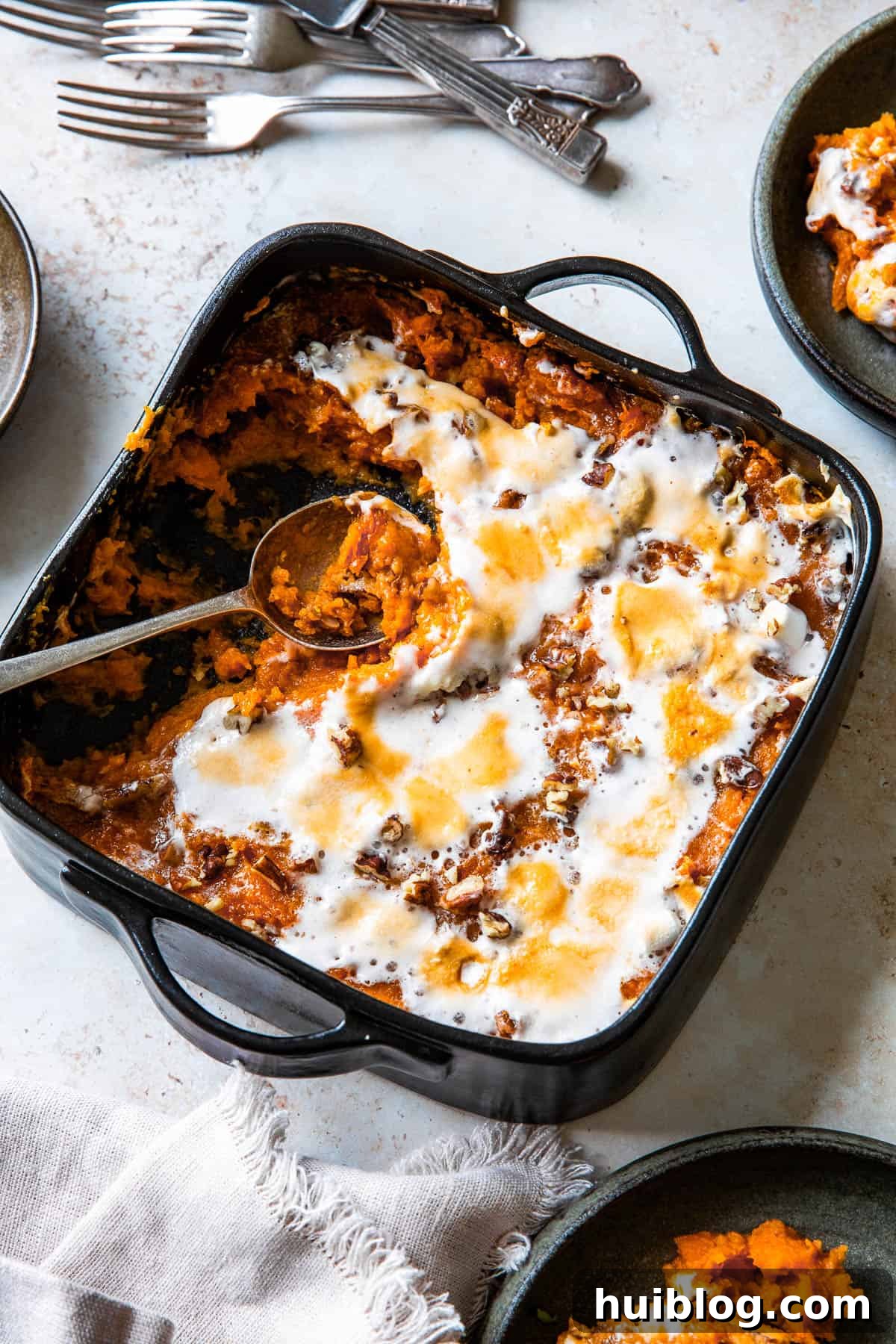 a square black baking dish filled with sweet potato casserole, topped with melted marshmallows. The marshmallow topping is golden-brown from baking. Several portions have been scooped out, and a serving spoon rests in the empty part of the dish.