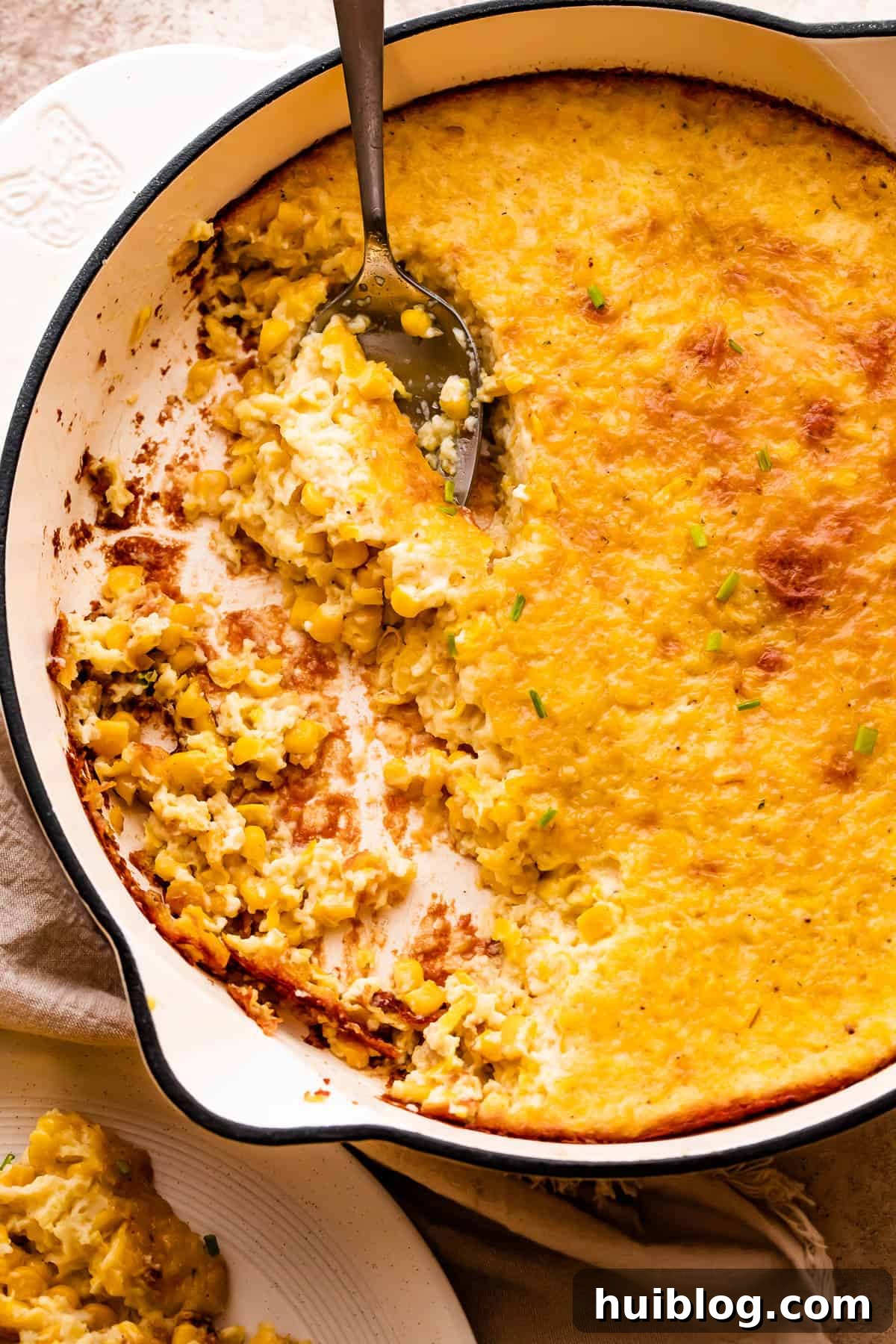 A spoon lifting a serving of golden-brown creamed corn pudding from a white cast iron skillet, showcasing its creamy texture.