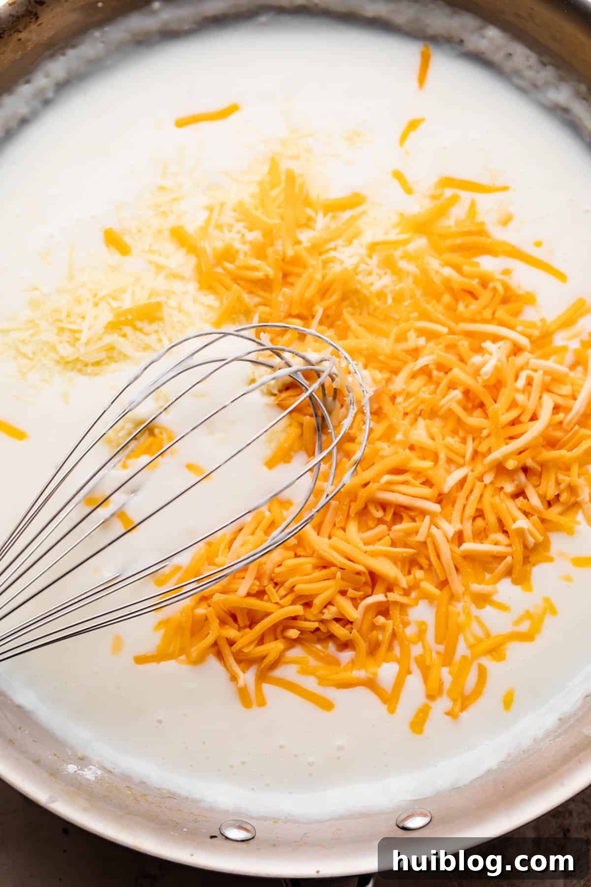 A close-up shot of a whisk combining shredded cheddar cheese with milk in a skillet, creating a smooth sauce.