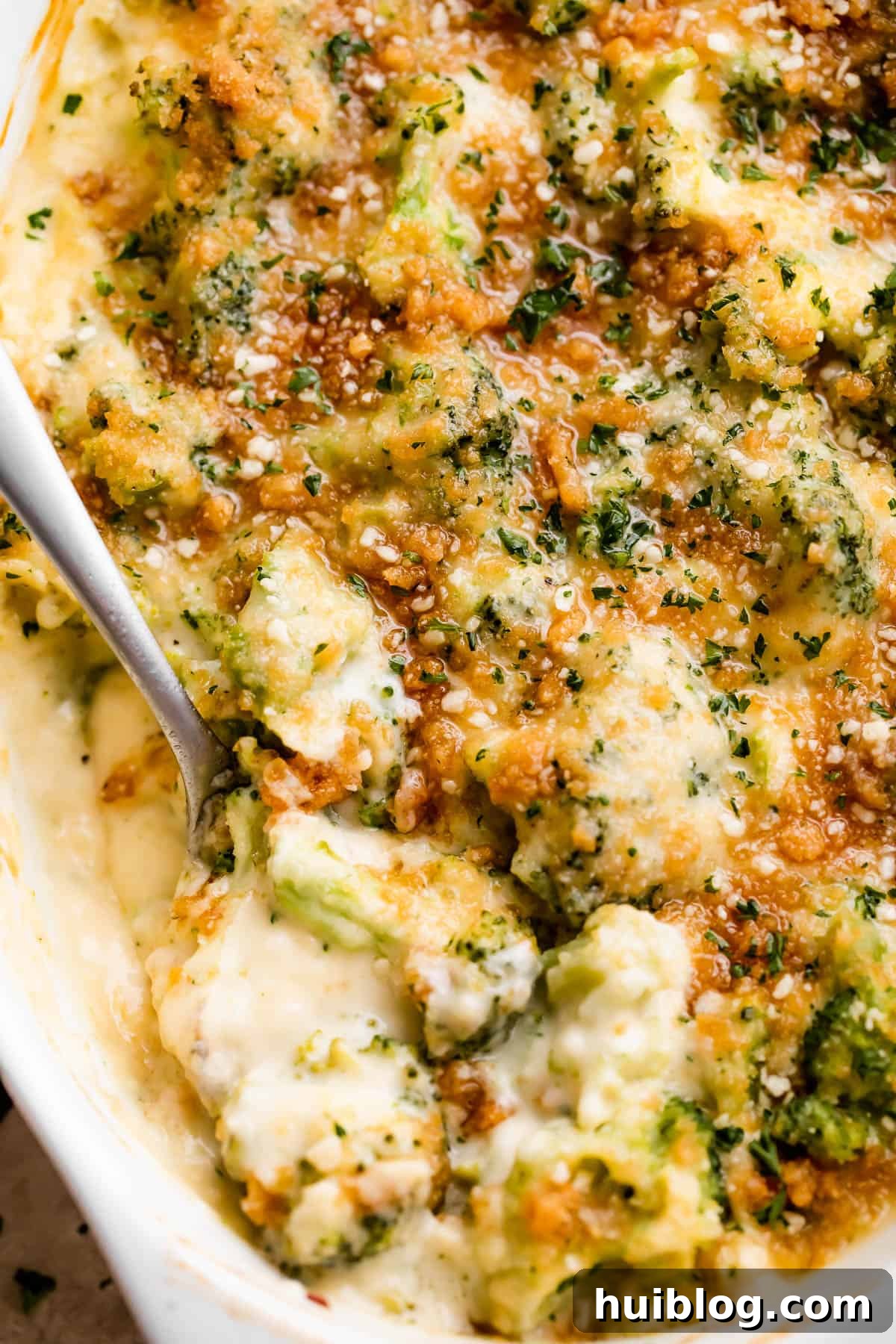 Overhead shot of an oval baking dish with Creamy, Cheesy Broccoli Casserole, a spoon mixing through its rich contents, ready to be served.