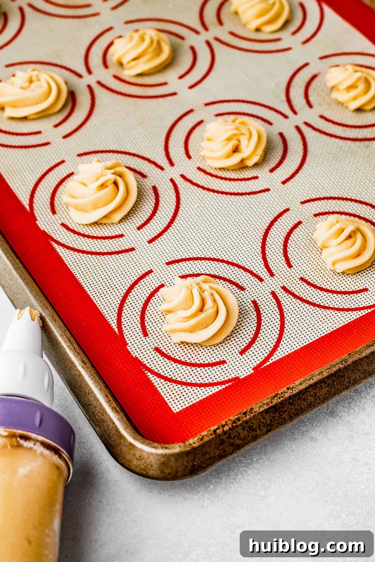 Rows of unbaked butter cookies, meticulously piped into elegant swirls on a silicone baking mat atop a large cookie sheet, ready for chilling and baking.