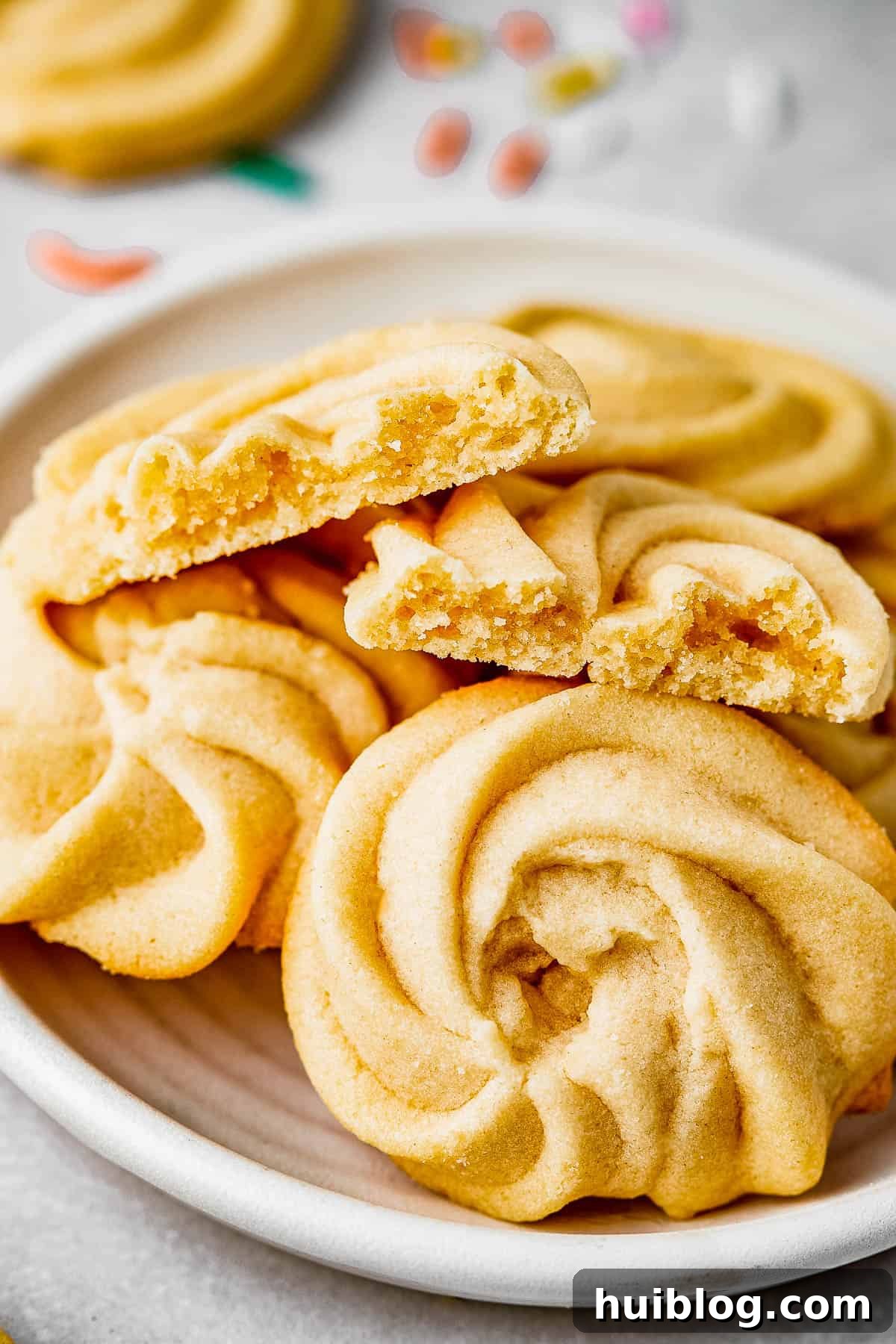 A close-up shot of four perfectly baked butter cookies arranged on a small plate. One cookie is broken in half, revealing its tender, crumbly interior and confirming its delicate texture.