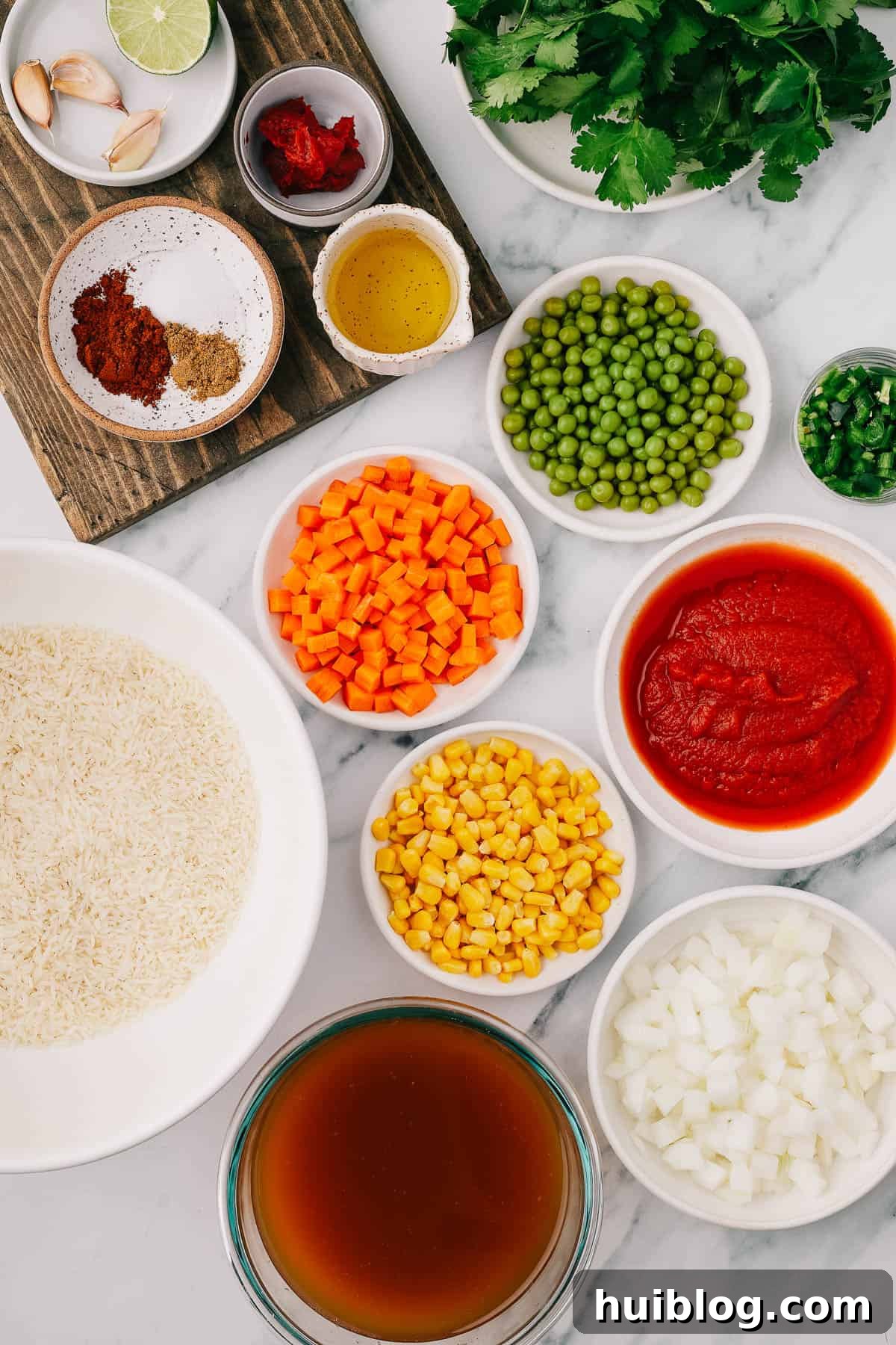 A selection of fresh and pantry ingredients for Instant Pot Mexican Rice, including garlic, lime, tomato paste, cilantro, chili powder, salt, cumin, oil, peas, jalapeno, rice, carrots, corn, tomato puree, vegetable broth, and onion, arranged neatly.