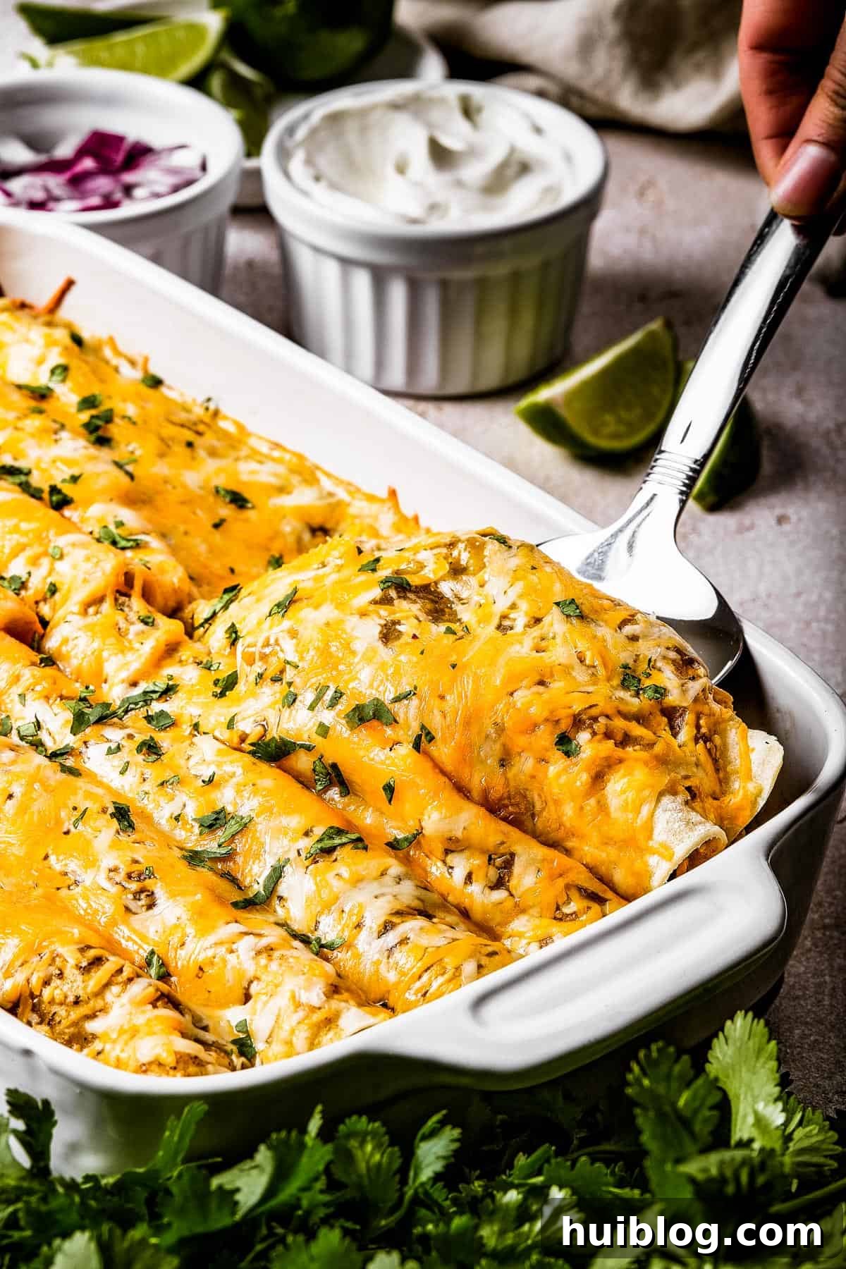A silver serving spoon lifting two cheesy salsa verde chicken enchiladas from a white baking dish, showcasing the melted cheese and green sauce.
