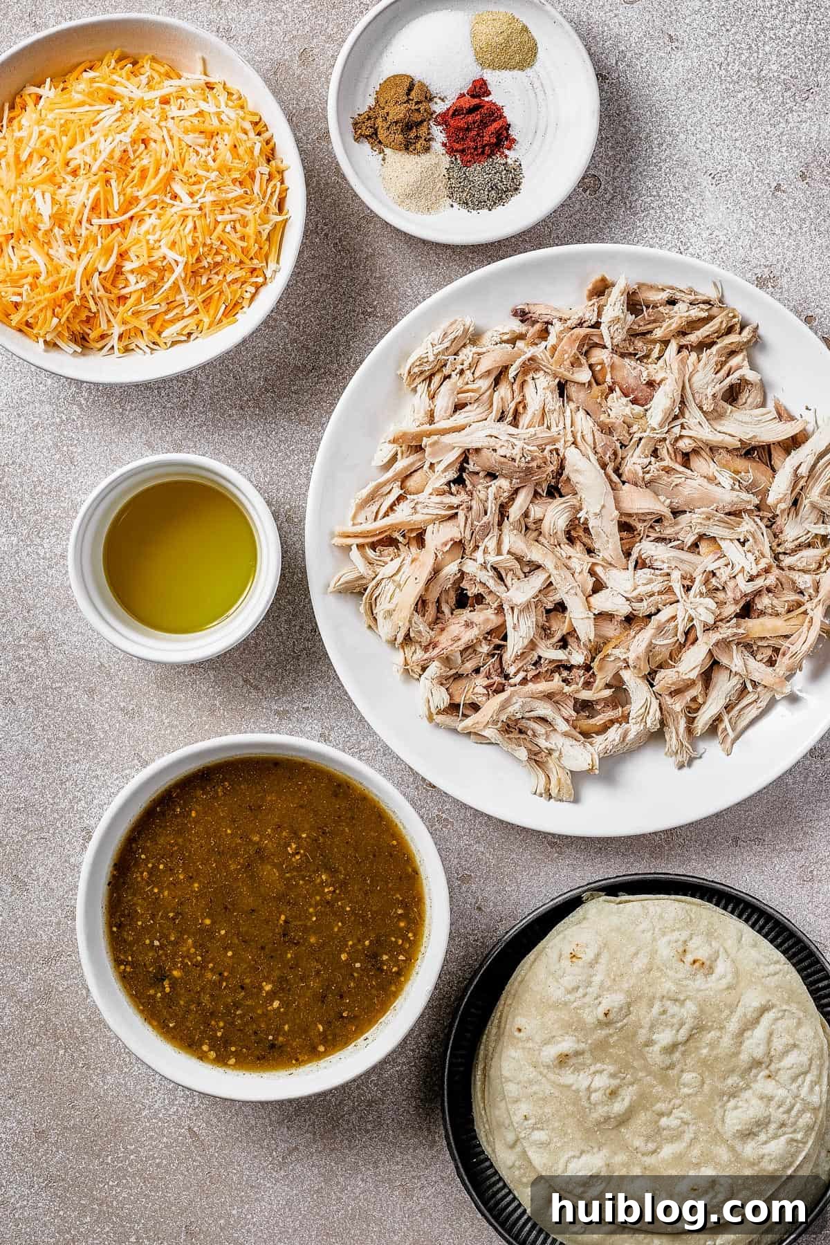 A flat lay photograph of all the core ingredients for salsa verde chicken enchiladas, arranged clockwise from top left: shredded Mexican blend cheese, a small bowl of mixed spices (cumin, paprika, garlic powder, onion powder), shredded rotisserie chicken, a stack of corn tortillas, a jar of salsa verde, and a bottle of olive oil.