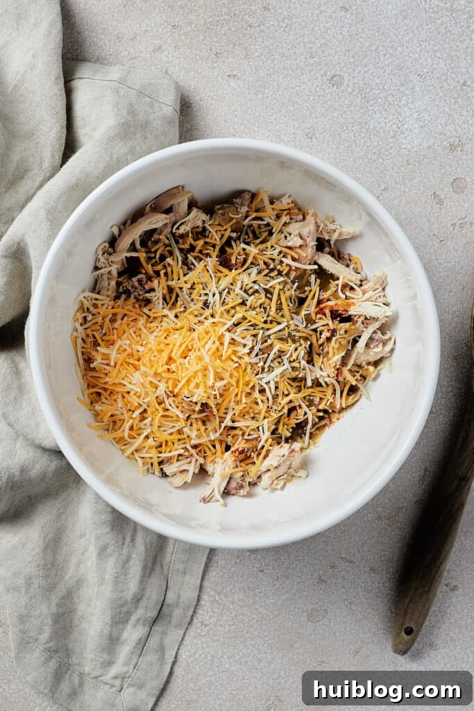 A white mixing bowl filled with shredded chicken, spices, and shredded cheese, ready to be mixed for the enchilada filling.