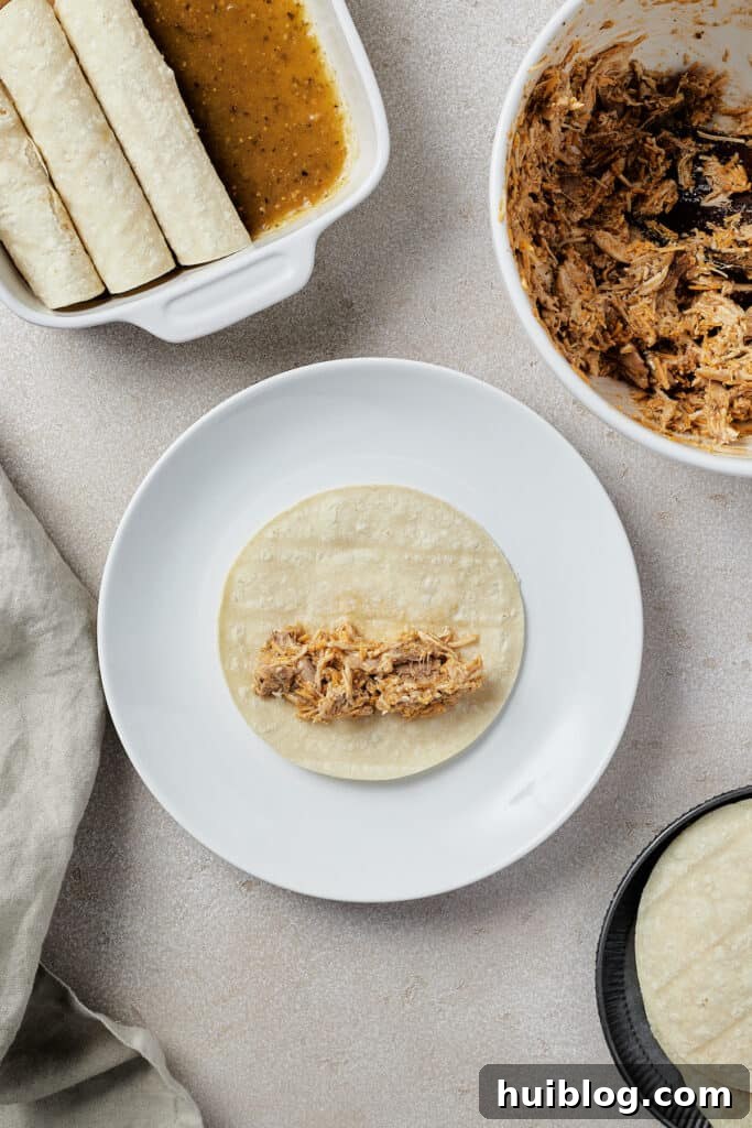 A small corn tortilla on a white plate with a spoonful of chicken and cheese filling placed in the center, ready to be rolled. In the background, a full baking dish of enchiladas, a bowl of filling, and more tortillas are visible.