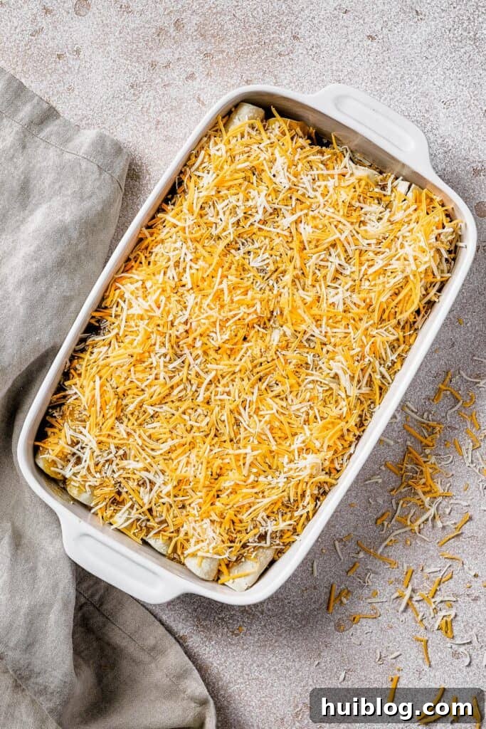 A white baking dish filled with rolled enchiladas, completely covered with shredded cheese, ready to be baked.