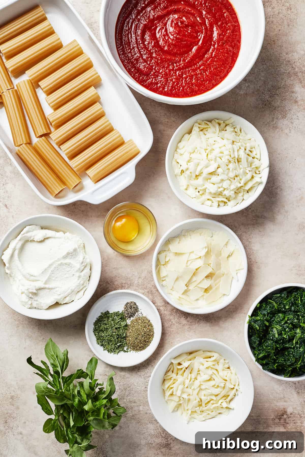 A flat lay photograph displaying all the essential ingredients for classic cheese manicotti: uncooked pasta shells, a jar of marinara sauce, various shredded cheeses (mozzarella, provolone, parmesan), fresh spinach, and herbs, alongside a single egg.