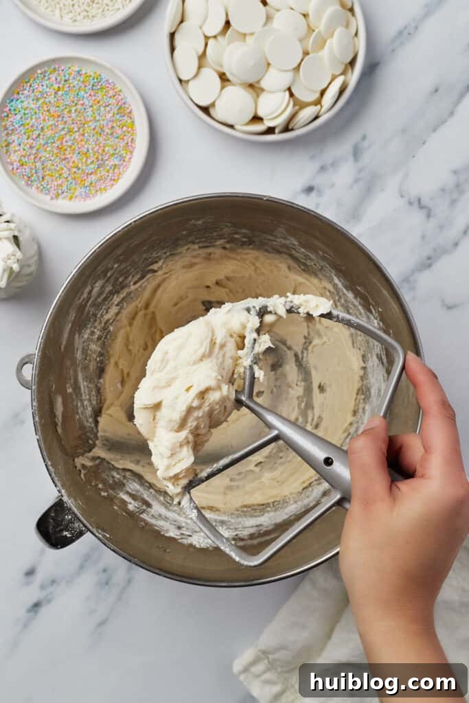 Creamy frosting in a mixing bowl, with a woman's hand lifting a stand mixer attachment covered with the delicious mixture.