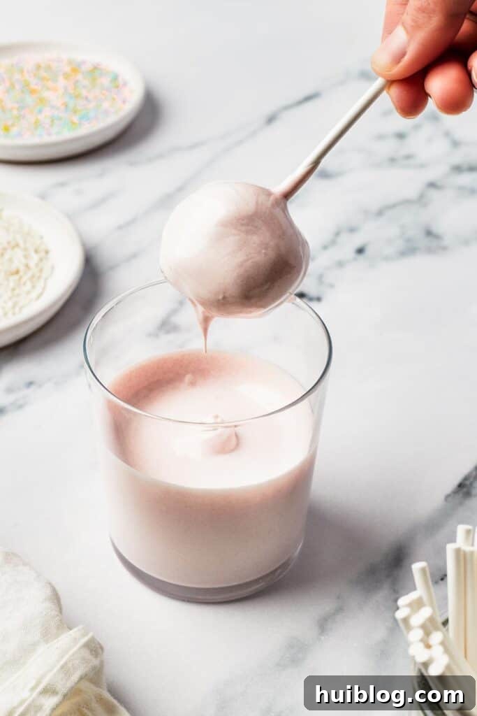 A cake pop ball being carefully dipped into a tall glass of melted candy coating, showing the precise moment of transformation.