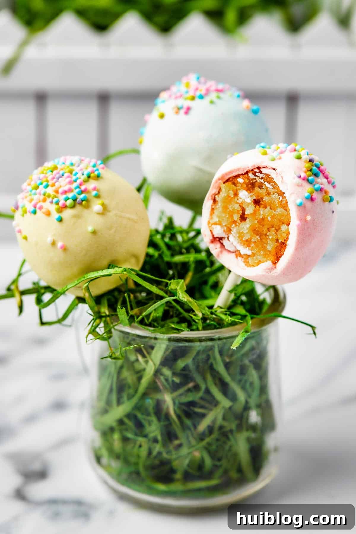 Three beautifully decorated cake pops in a small glass filled with Easter grass, one having a tempting bite taken out of it.