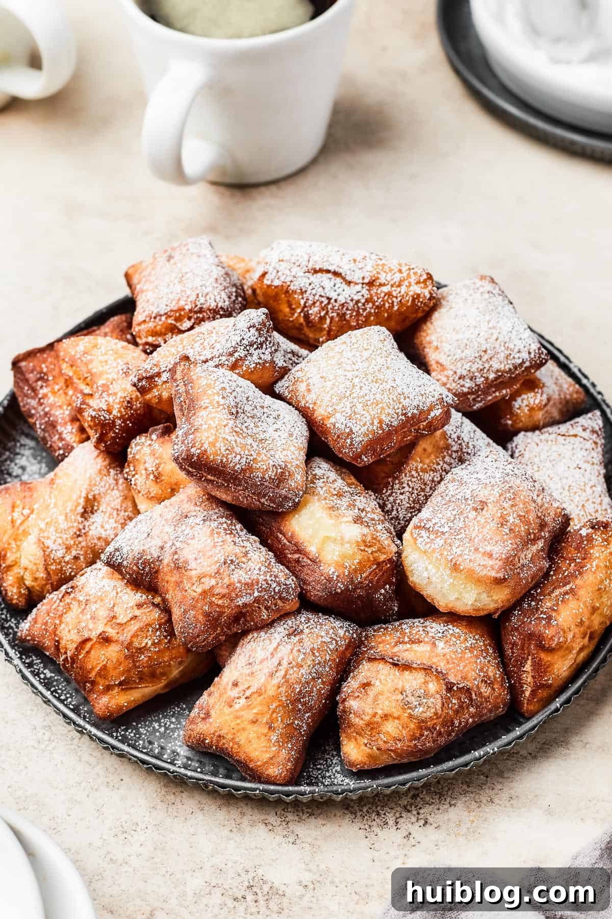 French Quarter Cloud Cakes 2 A plate of golden-brown, square beignets, generously dusted with powdered sugar, ready to be enjoyed.