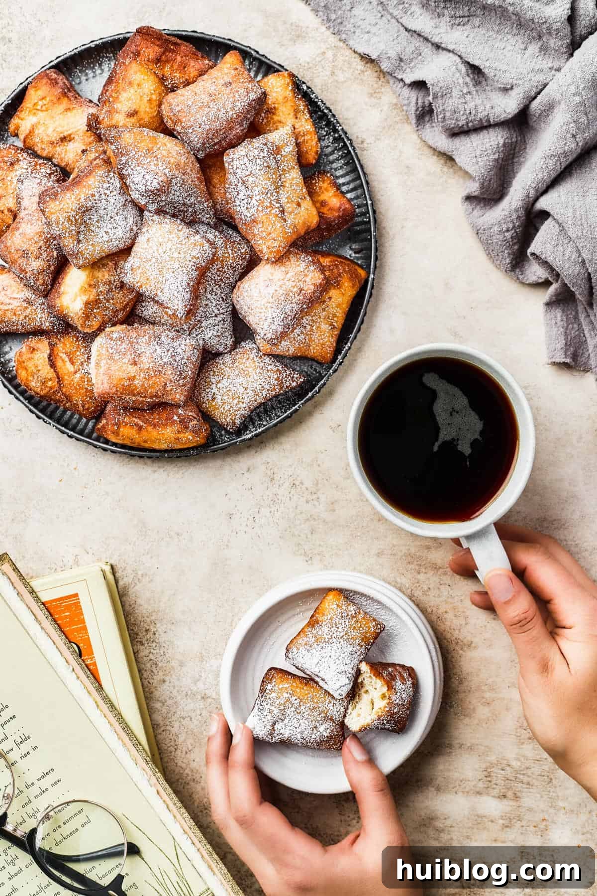French Quarter Cloud Cakes 3 An inviting overhead shot of a large platter overflowing with freshly fried and sugared beignets. A cup of dark coffee sits nearby, perfect for dipping.