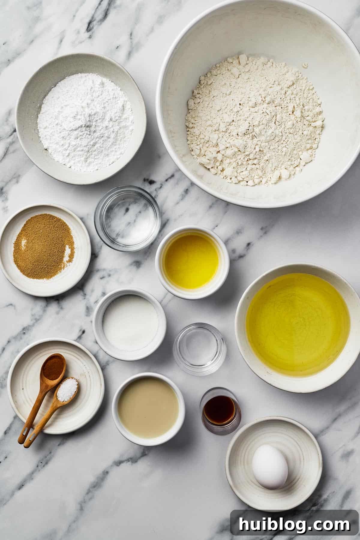French Quarter Cloud Cakes 4 A flat lay photograph showcasing all the essential ingredients needed for making beignets: powdered sugar, all-purpose flour, active dry yeast, warm water, melted unsalted butter, canola oil, granulated sugar, white vinegar, ground cinnamon, kosher salt, pure vanilla extract, and one whole egg.