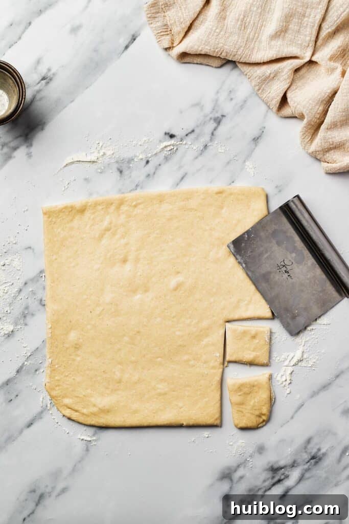 French Quarter Cloud Cakes 7 Squares of rolled-out yeast dough, freshly cut, sit on a marble work surface, ready for frying. A bench scraper rests nearby.