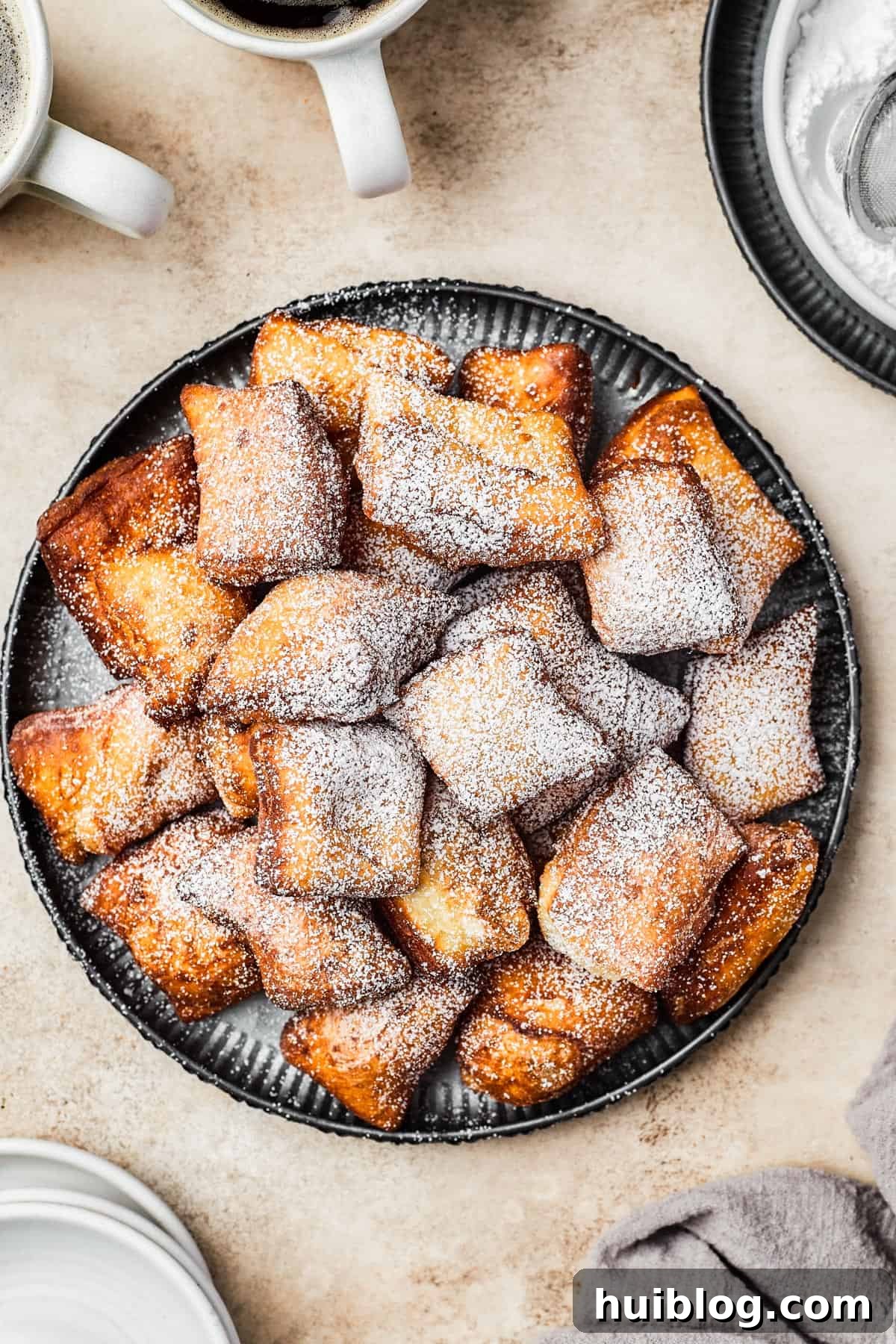 French Quarter Cloud Cakes 9 A freshly prepared plate of golden-brown fried beignets, piled high and generously coated with a delicate layer of powdered sugar.