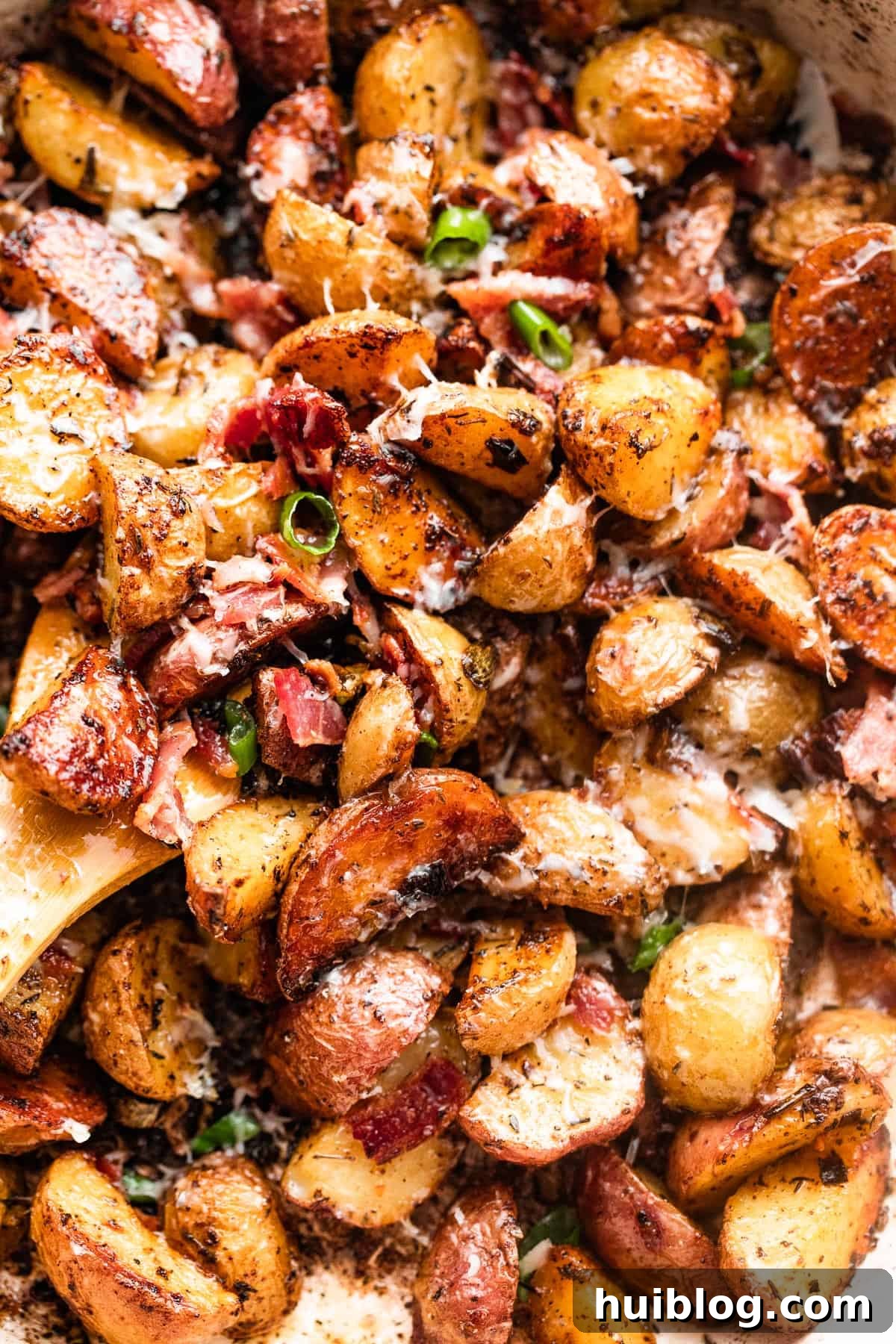 close up shot of a wooden spoon stirring through roasted potatoes with crispy bacon and melted Parmesan cheese.