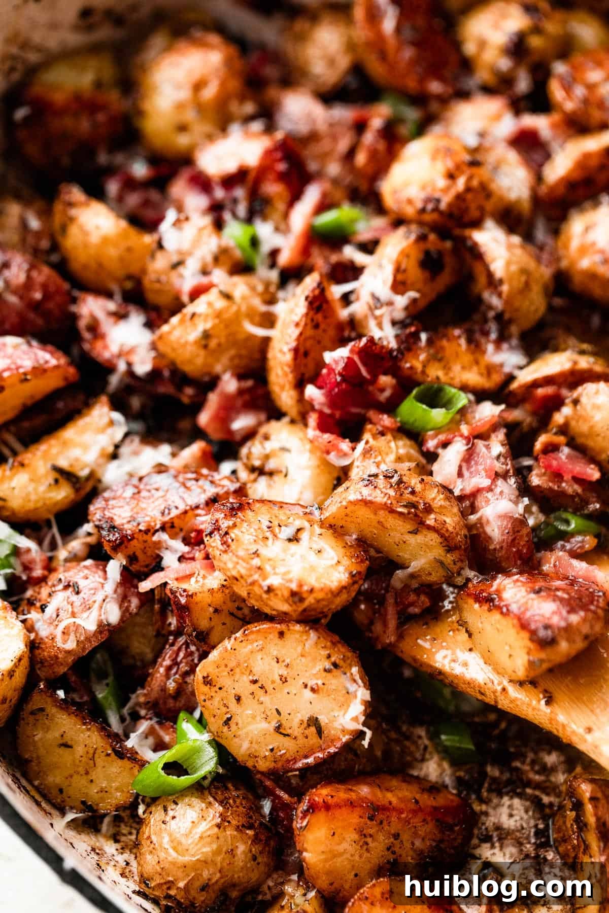 close up shot of a wooden spoon stirring through roasted potatoes with bacon and cheese, highlighting the delicious texture.
