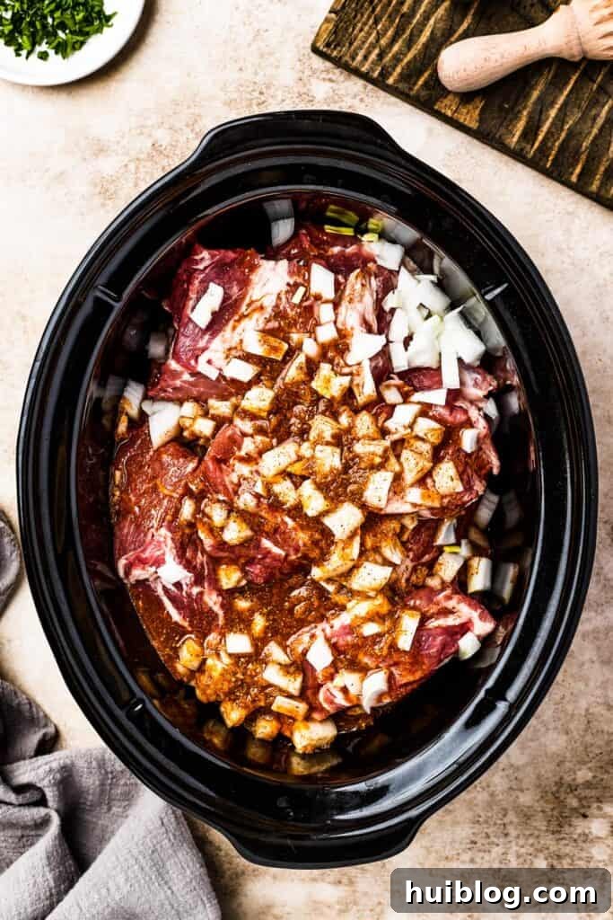 Chunks of boneless pork butt and diced white onion submerged in a rich, spiced liquid within a slow cooker.