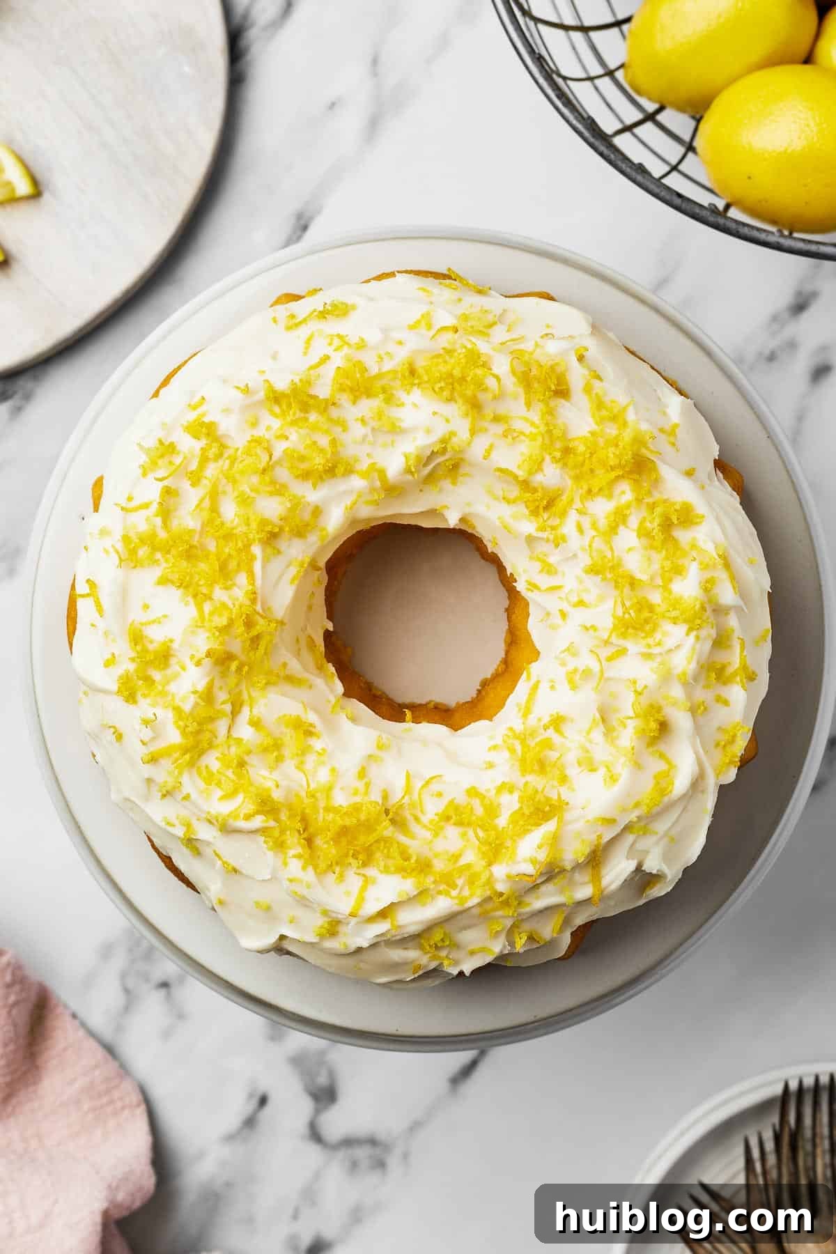A beautifully decorated lemon Bundt cake featuring smooth white frosting and a generous scattering of bright lemon zest, elegantly presented on a round white plate.