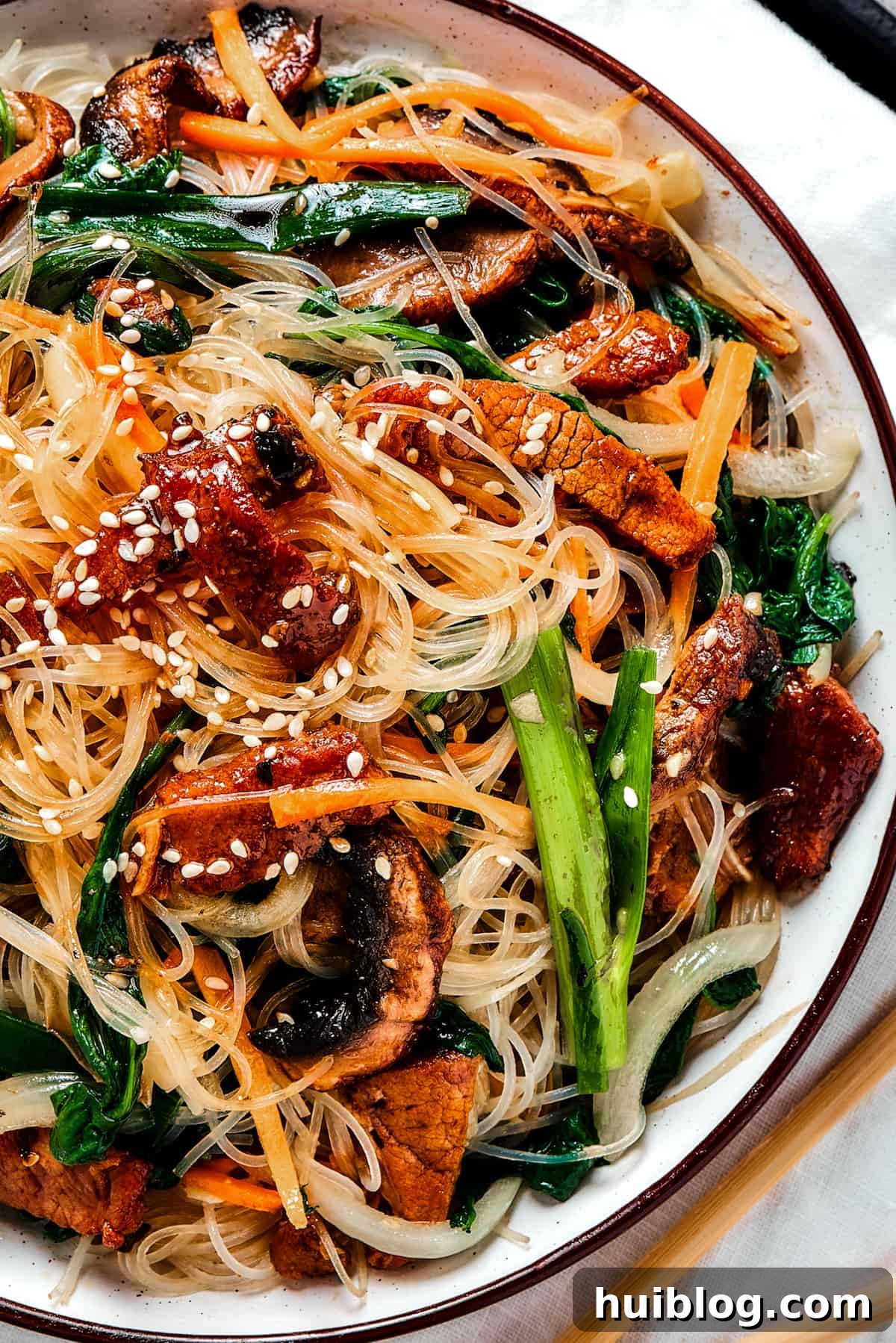 Authentic Korean Beef Japchae: A Delicious Glass Noodle Stir Fry for All Occasions Overhead shot of a shallow dish of beef Japchae (Korean glass noodle stir fry), garnished with toasted sesame seeds.