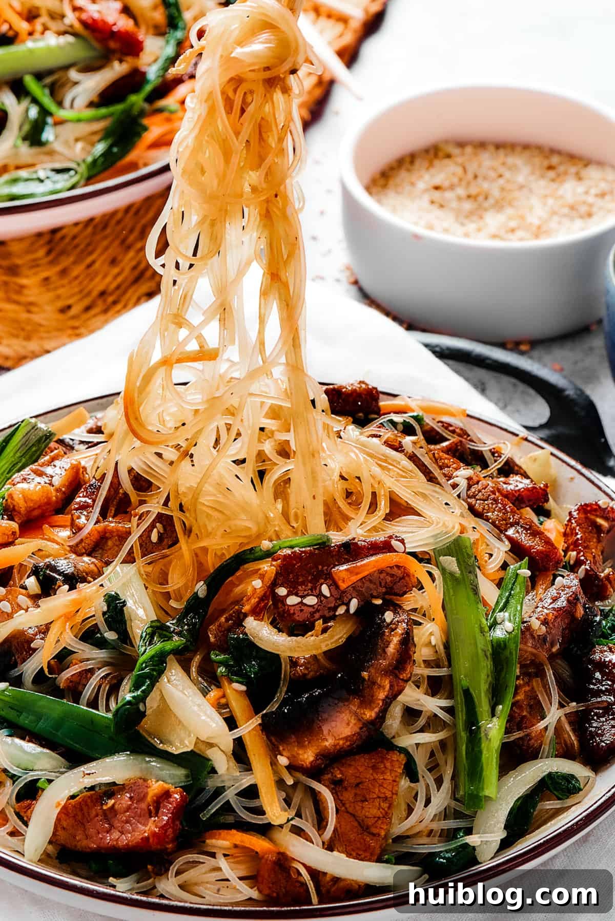 The Enjoyment of Japchae A bowl of Korean Beef Japchae with glass noodles. Some of the noodles are being lifted from the bowl with chopsticks, showing their appealing texture.