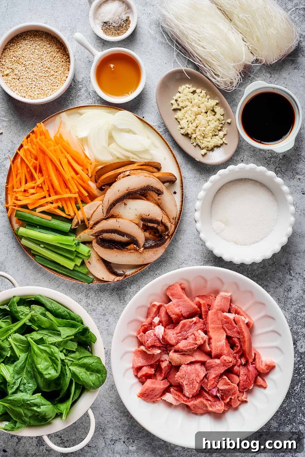 Essential Ingredients for Authentic Korean Beef Japchae A vibrant flat lay showcasing all the fresh ingredients for Korean Beef Japchae: toasted sesame seeds, salt and pepper, dried glass noodles, sesame oil, garlic, soy sauce, carrots, onions, green onions, mushrooms, sugar, spinach, and thinly sliced steak.