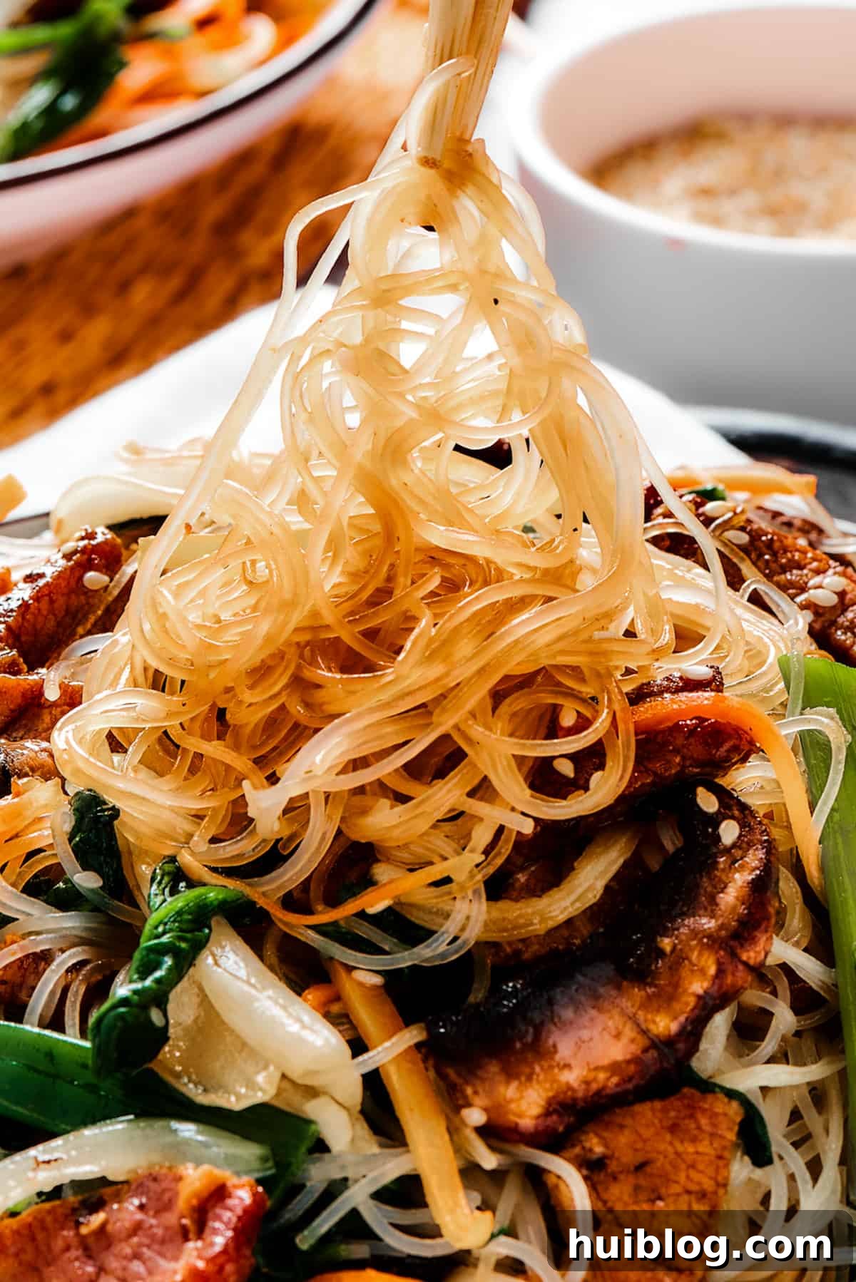 Savoring the Perfect Plate of Japchae A bowl of vibrant Korean Beef Japchae with glass noodles. Some of the springy noodles are being lifted from the bowl with chopsticks, highlighting the texture and rich sauce.