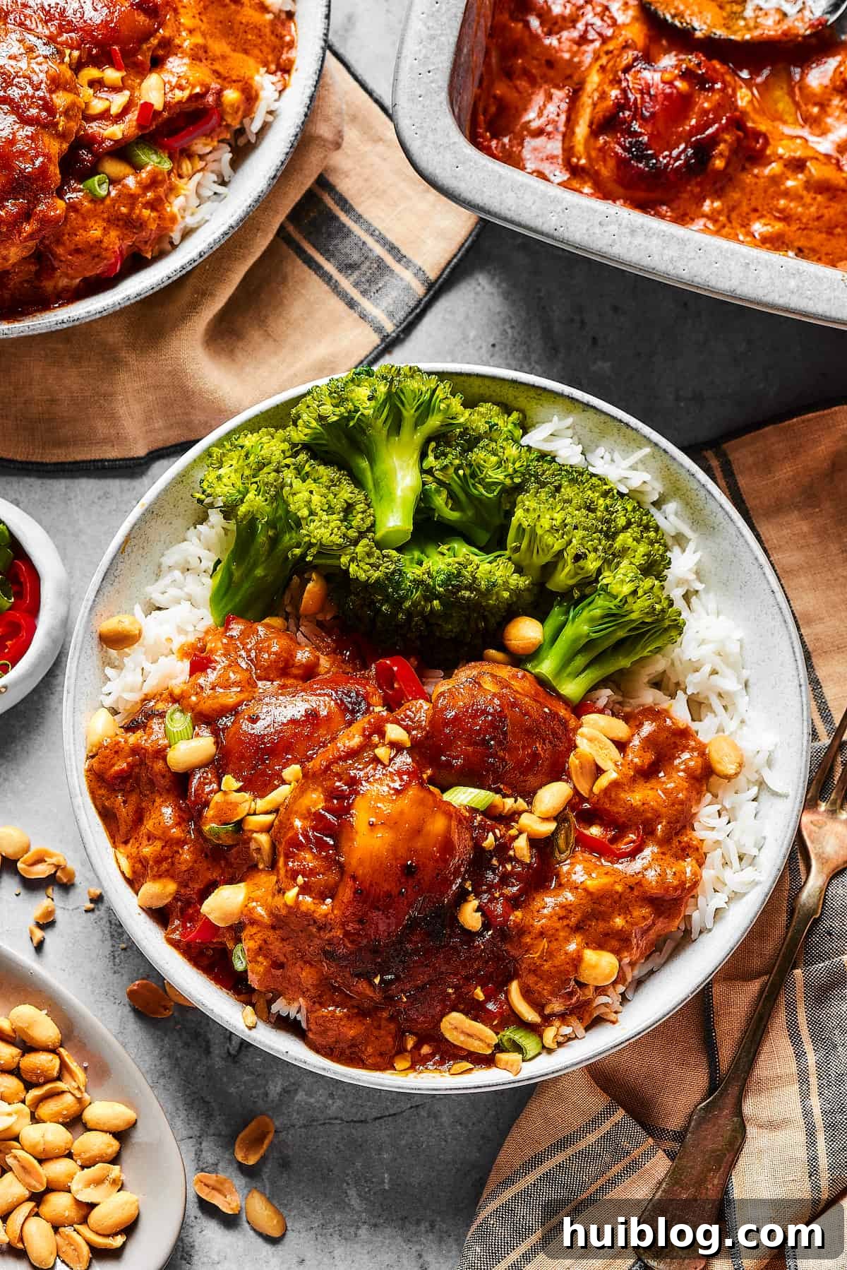 Simple Peanut Butter Chicken - Easy, Healthy, and Flavorful! Delicious Peanut Butter Chicken served alongside fresh broccoli and rice. A baking dish of the finished chicken, garnishes, and additional plates are visible on a rustic wooden table.