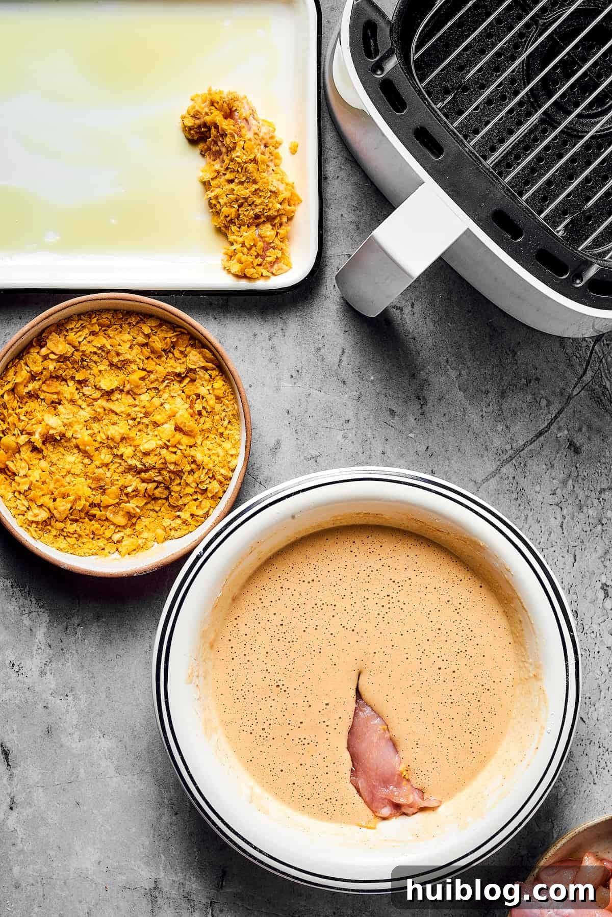 A piece of chicken dipped in batter, next to a bowl of cornflakes. A breaded chicken strip is on an oiled baking sheet nearby, along with an open air fryer, demonstrating the cooking process.