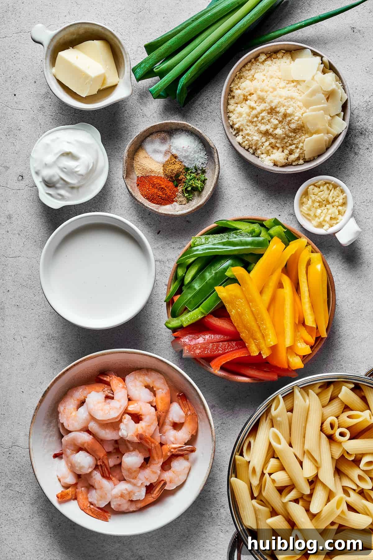 A flat lay photograph showcasing all the fresh ingredients for Rasta Pasta: butter, green onions, parmesan cheese, cream cheese, various spices, minced garlic, heavy cream, sliced red, yellow, and green bell peppers, raw shrimp, and uncooked penne pasta.