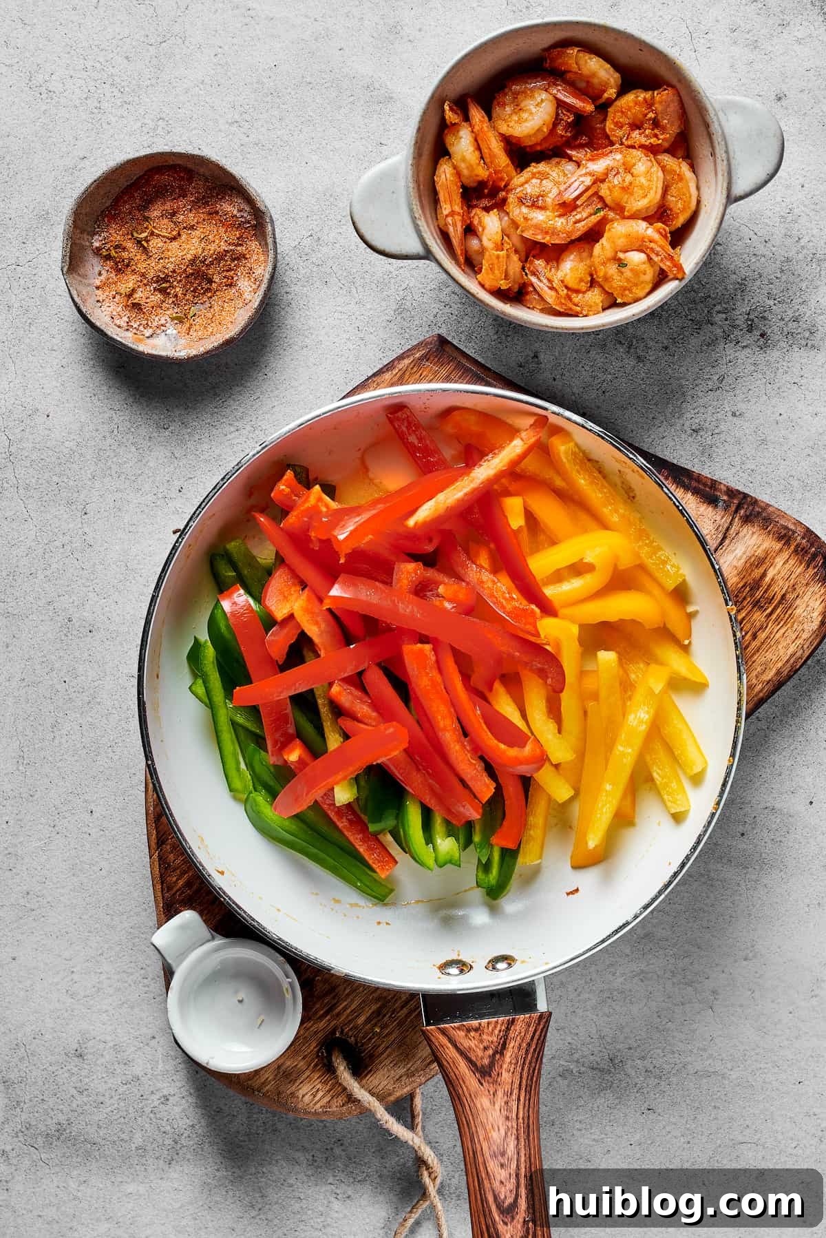 Green, red, and yellow bell peppers being sautéed in a skillet, next to a bowl of spicy marinated shrimp.