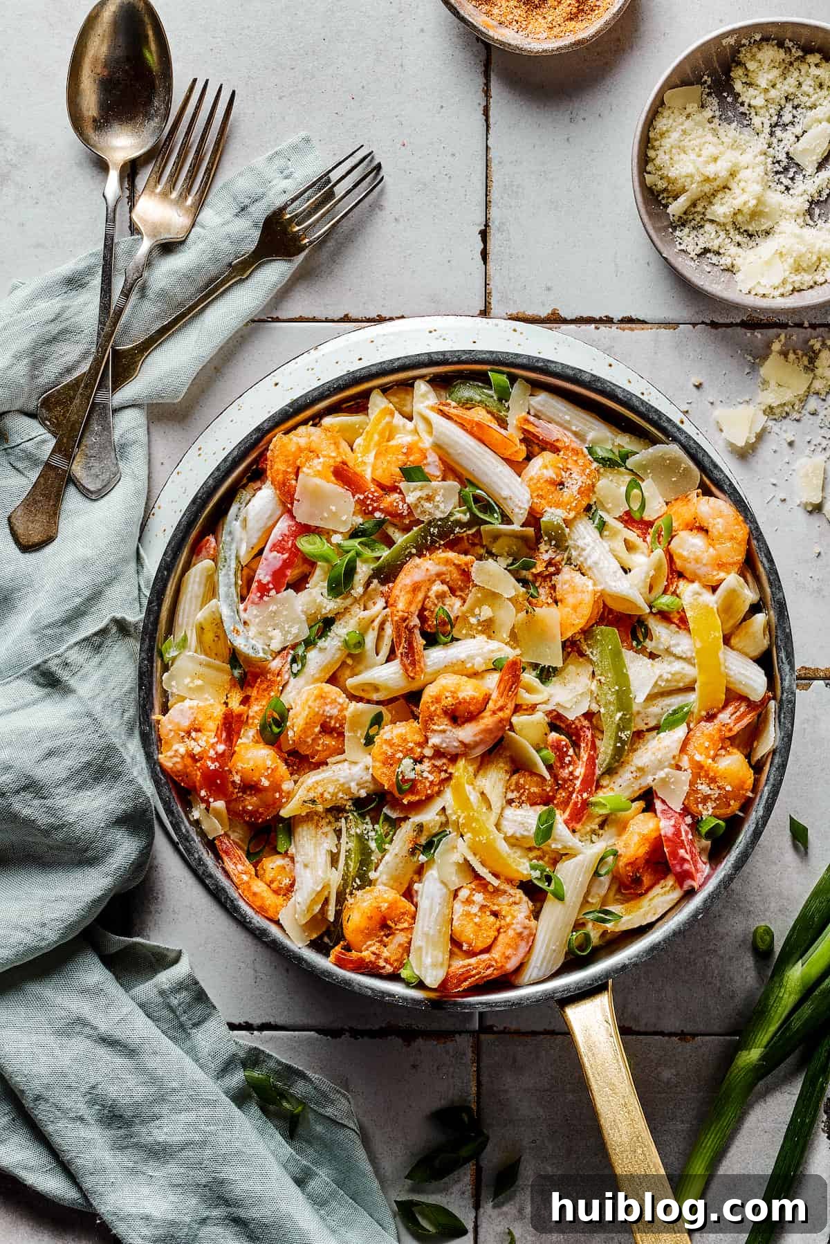 A complete skillet of Rasta pasta with serving spoons, forks, and a cloth napkin on a rustic surface.