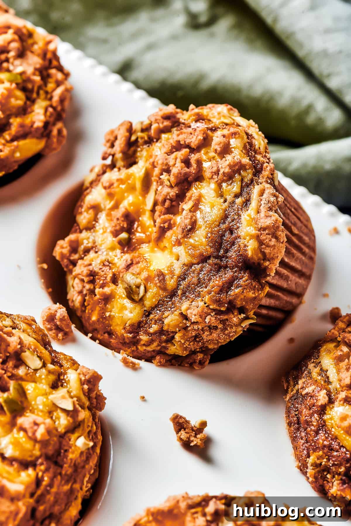 Pumpkin Cream Cheese Muffins are a heavenly mix of spiced pumpkin cake and smooth cream cheese swirl, with a buttery, streusel-like topping. A muffin tin with one pumpkin cream cheese muffin turned diagonally in its well, showcasing its golden streusel topping. Small pieces of topping have fallen off onto the muffin tin, hinting at its delightful texture.