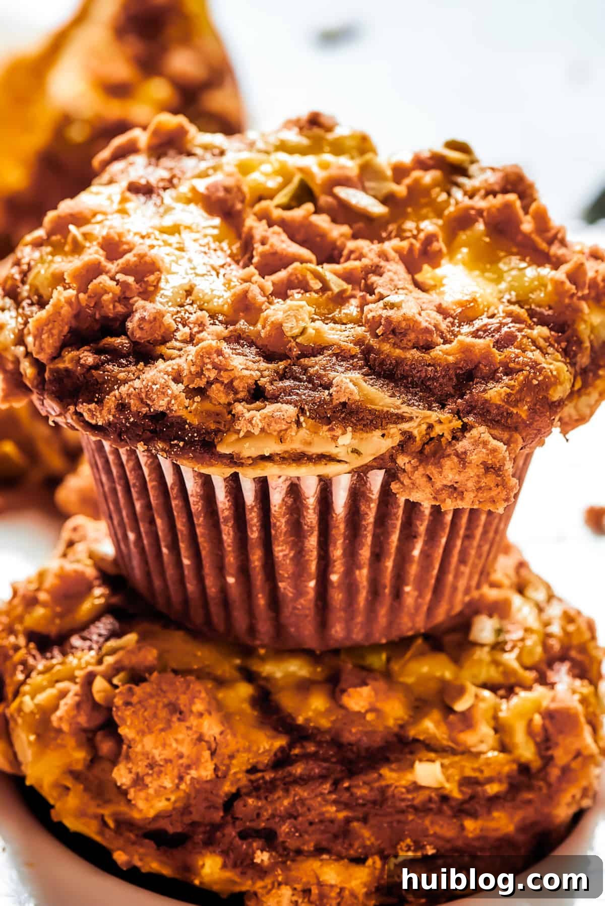 Pumpkin Cream Cheese Muffins are a heavenly mix of spiced pumpkin cake and smooth cream cheese swirl, with a buttery, streusel-like topping. A perfectly baked pumpkin cream cheese muffin set atop another muffin, arranged on a rustic wooden work surface.