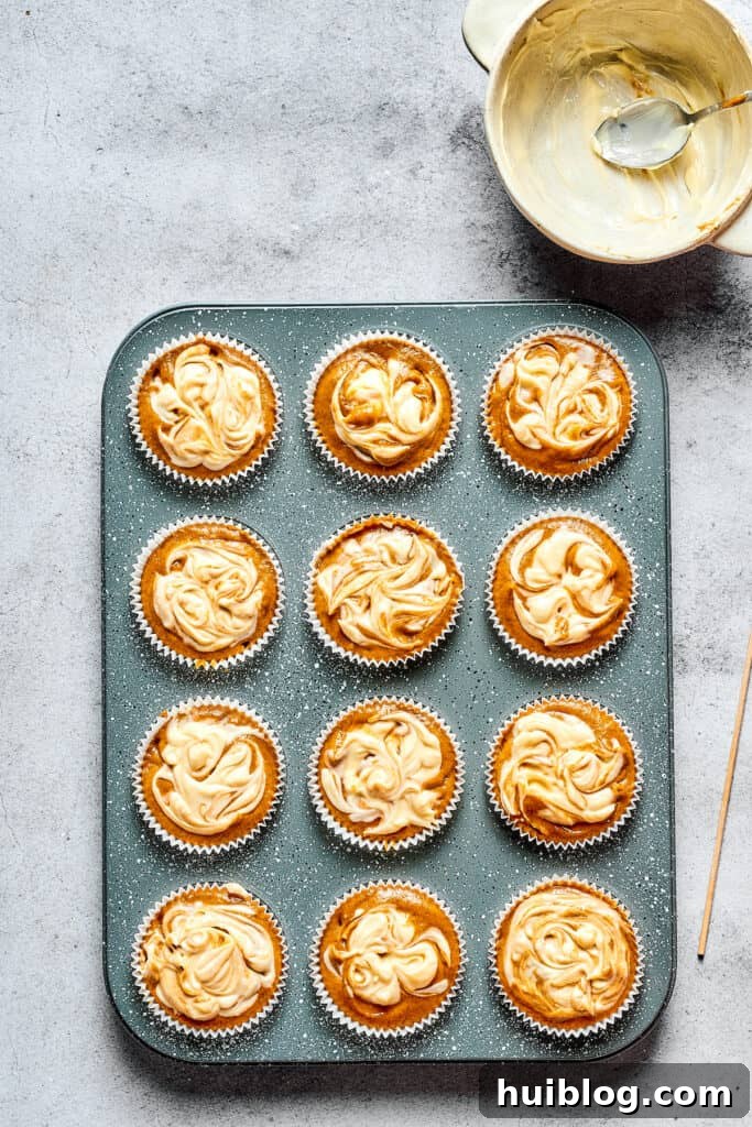 Swirled Pumpkin Cream Cheese Muffins 6 Unbaked pumpkin cream cheese muffins in a tin, each with a visible, piped cream cheese swirl ready for baking.