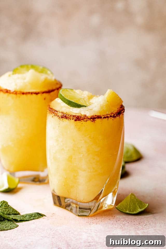 Two glasses of frozen pineapple margaritas, identical to the first image, showcasing their appealing presentation with chili-salt rims, lime, and mint.