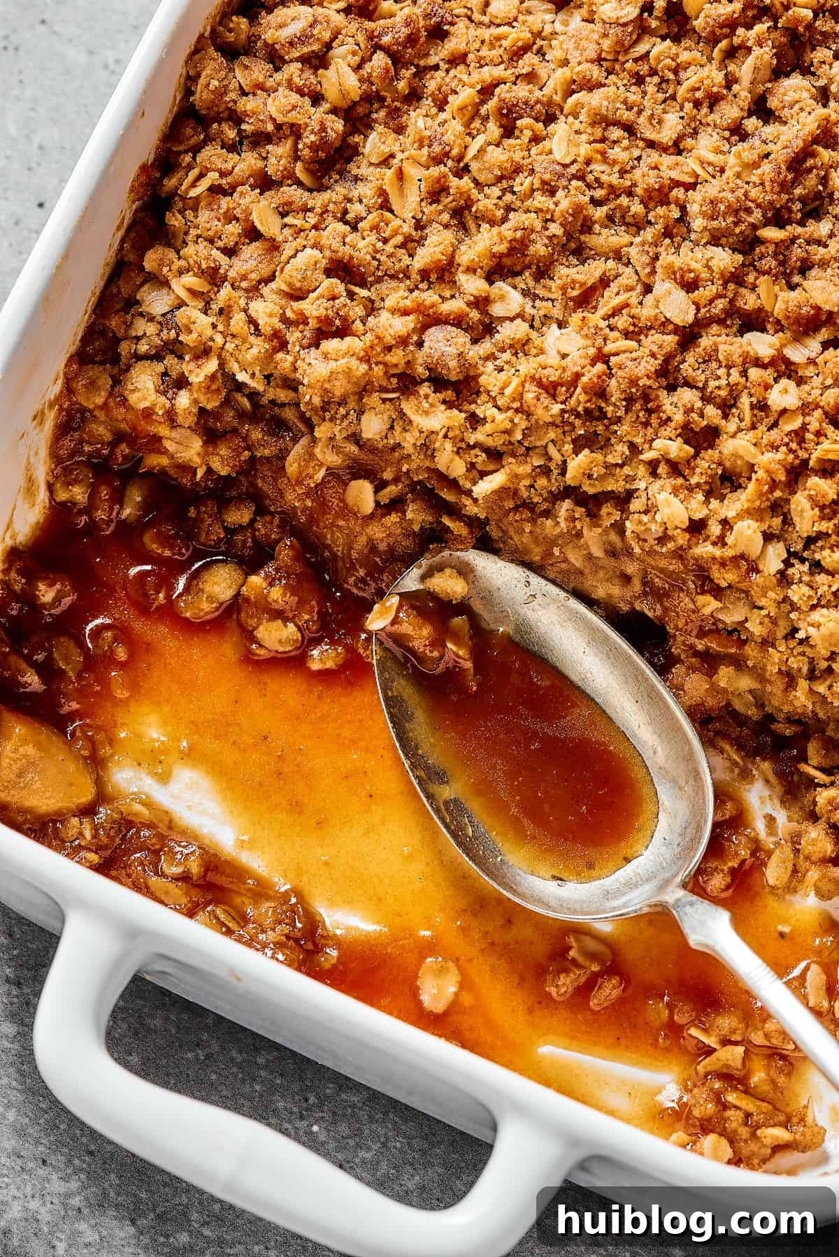 Close-up shot of the corner of a white casserole dish with an apple dessert, showing the apples, topping, and syrup. A spoon is resting in the empty part of the casserole dish.
