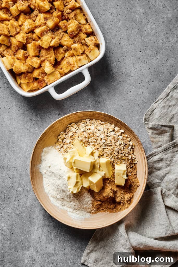 Spiced, floured apples in a casserole dish, next to a bowl of oats, cubed butter, and other baking ingredients.