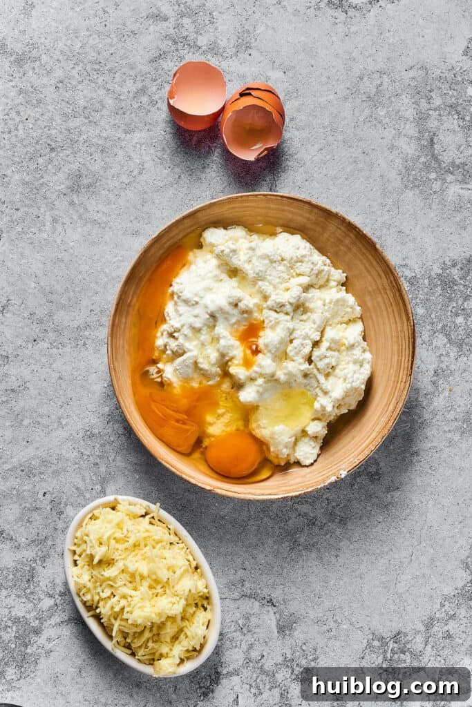 Cottage cheese, parmesan cheese, eggs, and other cheese filling ingredients in a bowl. A smaller bowl of shredded mozzarella is nearby on the work surface.