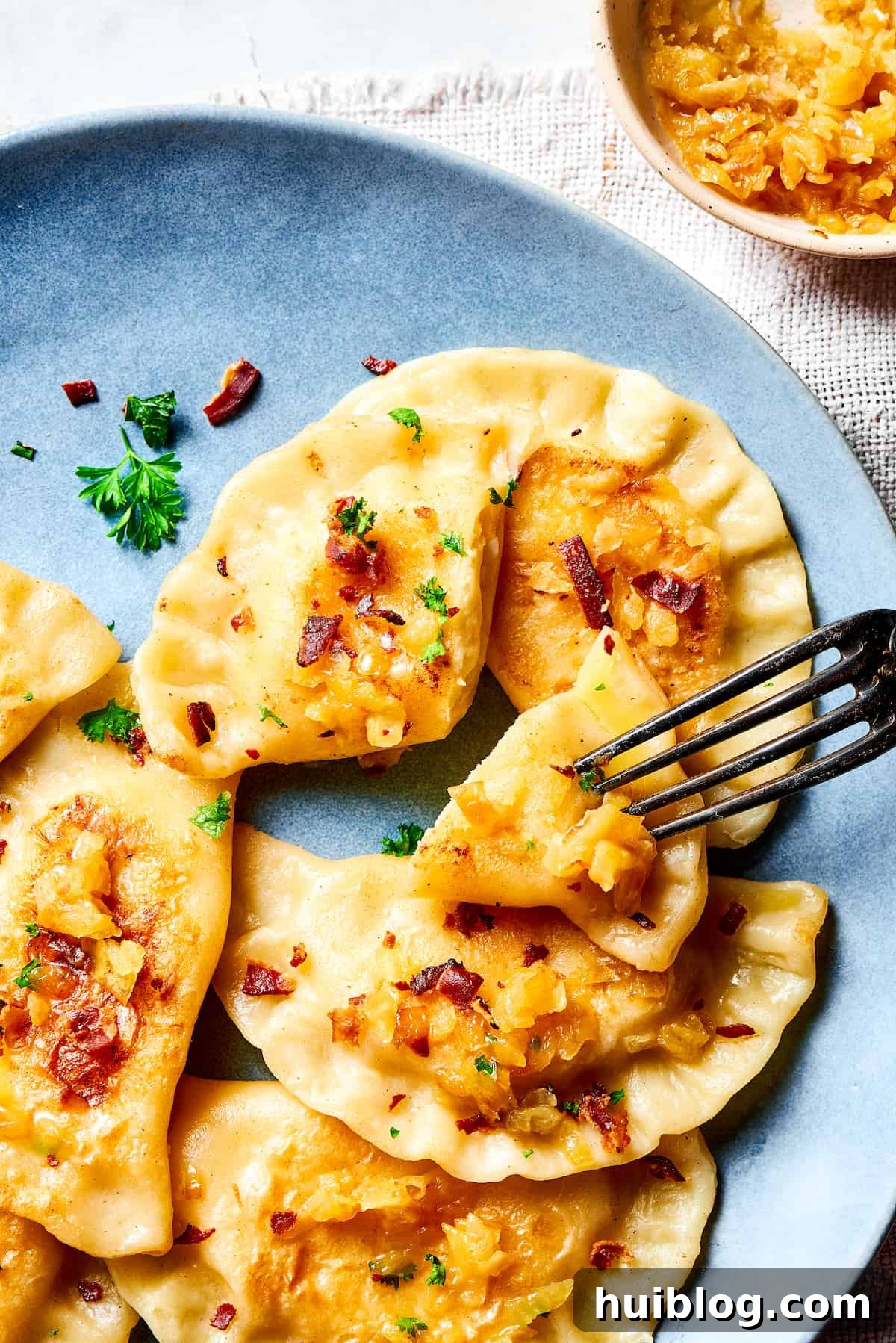 Topped with caramelized onion and crispy bacon, this easy Homemade Pierogi Recipe features a traditional cheese and potato filling. Yum! A fork piercing a bite-sized piece of a pierogi.