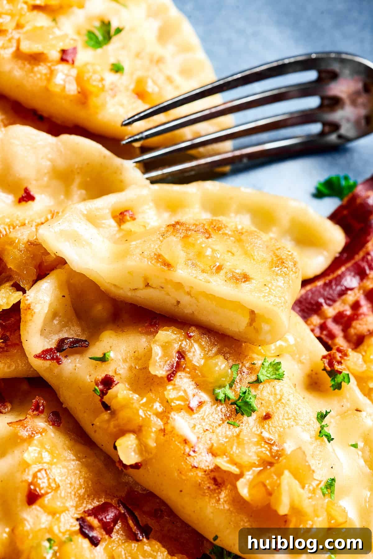 Topped with caramelized onion and crispy bacon, this easy Homemade Pierogi Recipe features a traditional cheese and potato filling. Yum! A plate of pierogies. One has been cut in half with a fork.